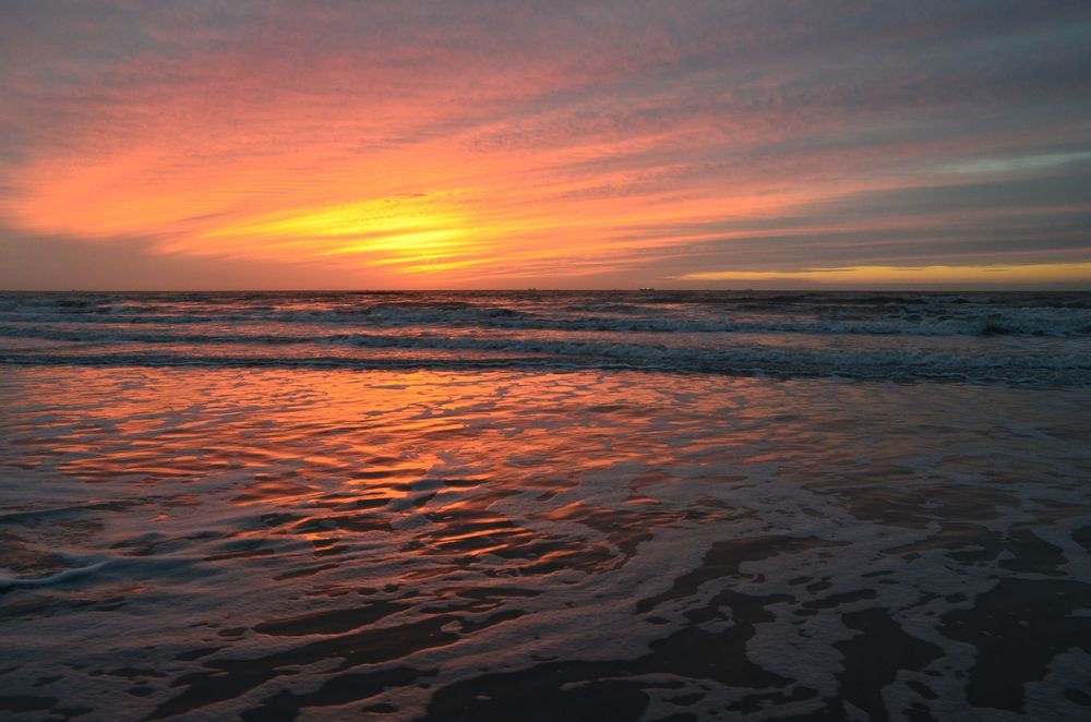 A glowing sunrise washes the shoreline in warm hues of orange, pink, and purple as gentle waves roll onto the beach