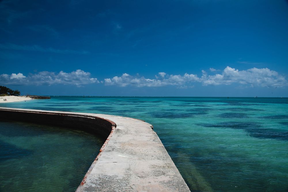 A curved concrete pier extends into vibrant turquoise water, surrounded by shifting blue‑green tones.