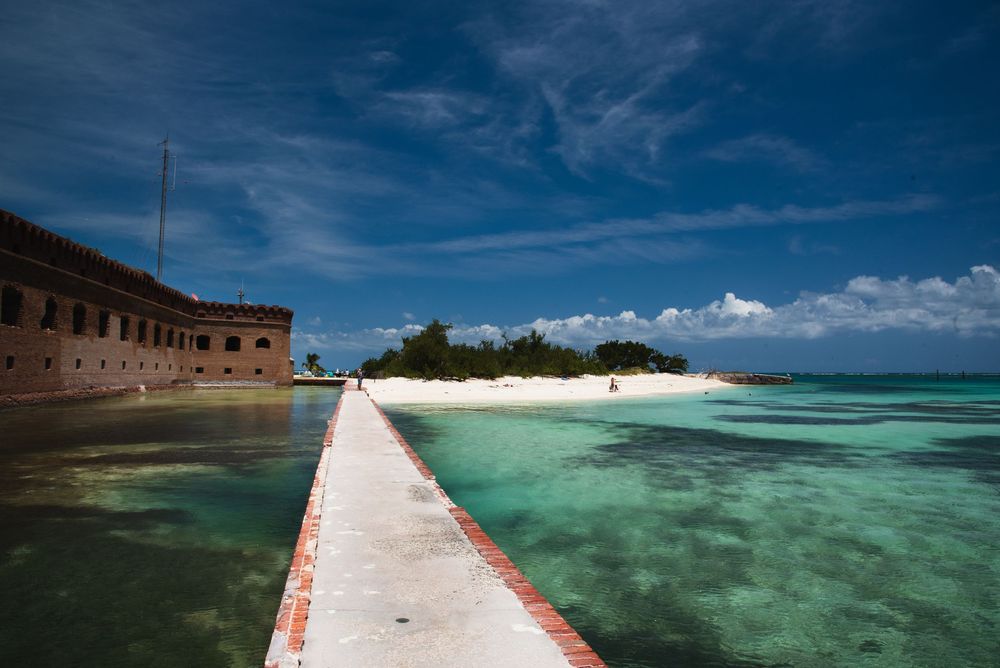 A historic brick fort overlooks a long, narrow walkway stretching across crystal‑clear turquoise water toward a small sandy island.