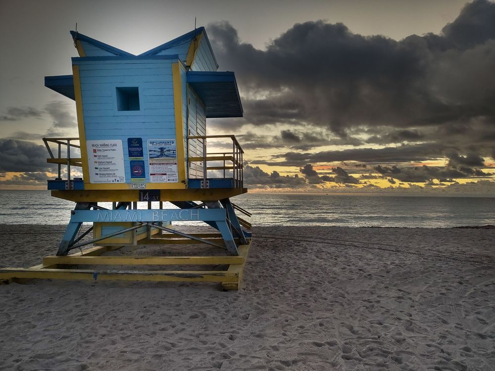 A colorful Miami Beach lifeguard tower stands against a dramatic sunrise sky, with warm light illuminating the sand and ocean.