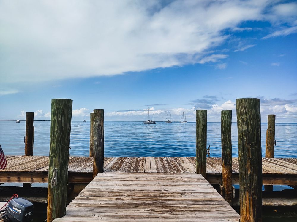 A weathered wooden dock stretches into calm blue water dotted with sailboats. Soft reflections ripple across the bay beneath a bright sky.