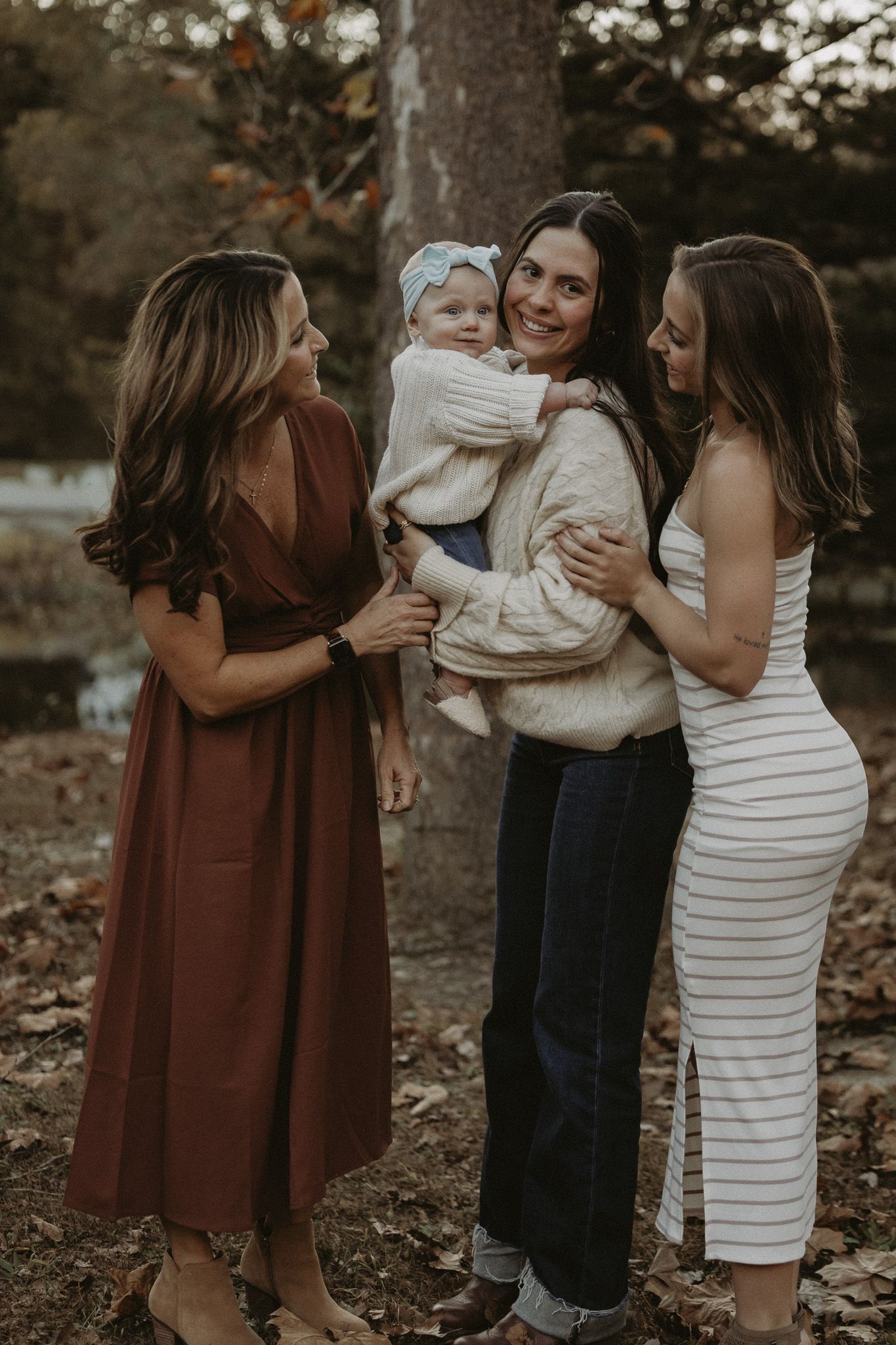 Three women smiling and gathering around a baby in cream sweater outdoors among autumn leaves.