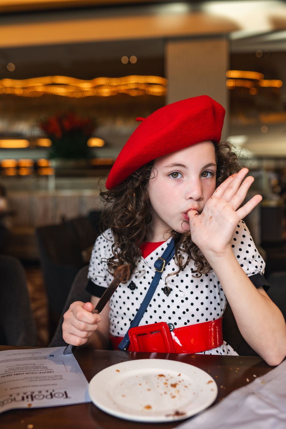 A person in a red beret and polka dot dress sits at a restaurant table with an empty plate.