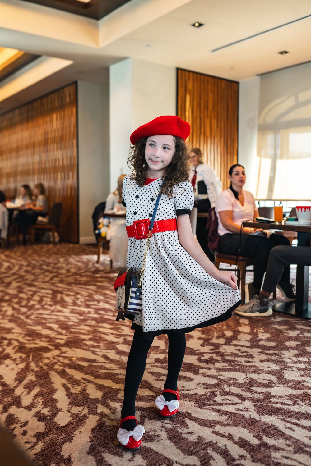 A young child in a polka dot dress and red beret twirls playfully on a patterned hotel carpet.