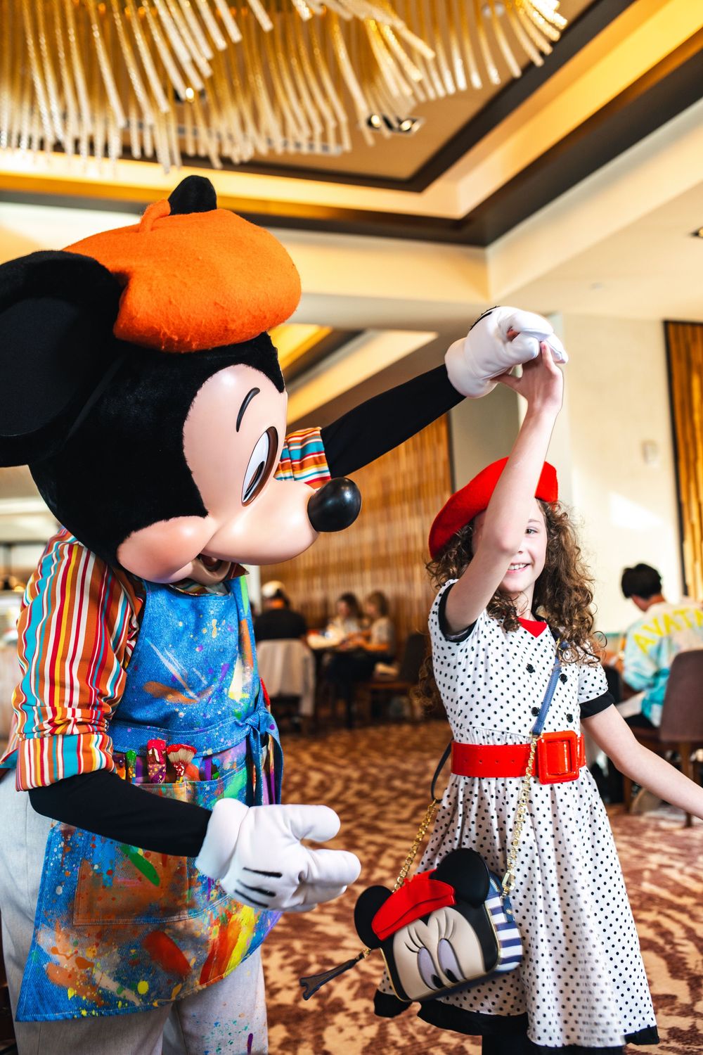 Mickey Mouse character in colorful outfit dances with young guest in polka dot dress at Disney restaurant.