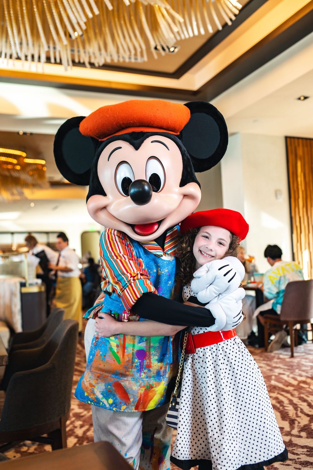 Mickey Mouse character hugs guests in a polka dot dress at a Disney meet and greet event in a hotel restaurant.