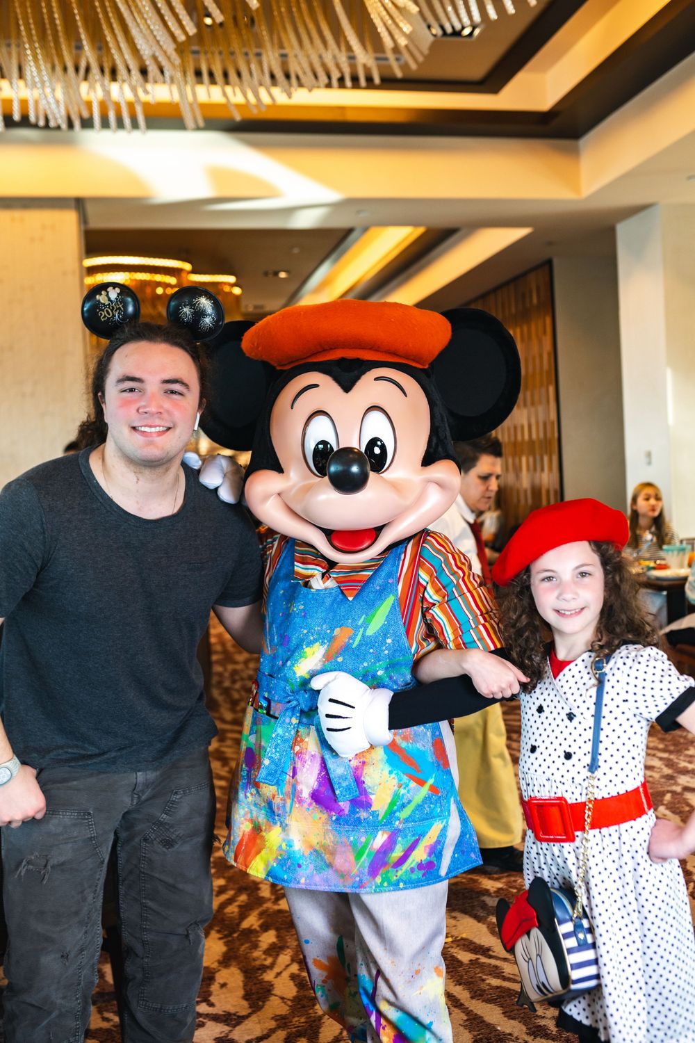 Mickey Mouse character posing with guests in polka dot dress and colorful tie-dye shirt at indoor Disney meet and greet.