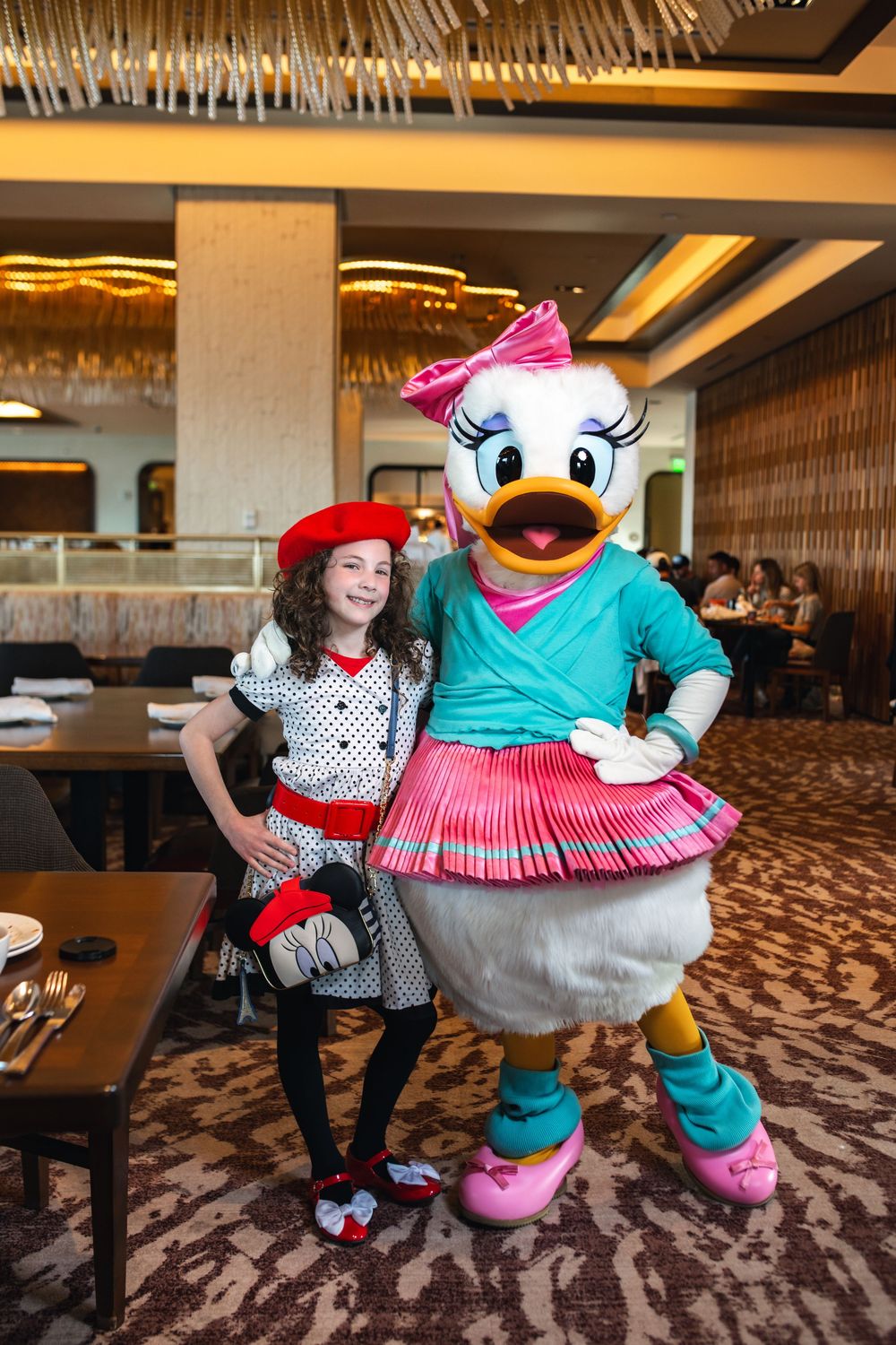 Daisy Duck character mascot poses with guest in polka dot dress at Disney hotel restaurant.