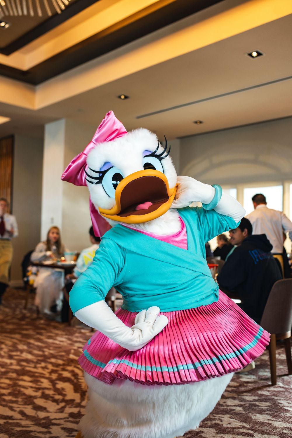 Daisy Duck character in turquoise top and pink pleated skirt performs animated pose at Disney dining experience.