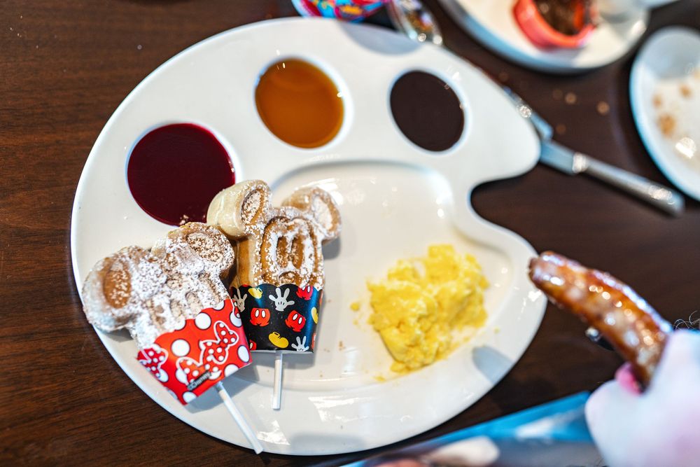 An artist's palette holds colorful paint sauces and condiments next to decorated cookies and yellow garnish on a wooden table.