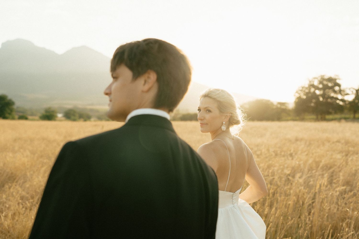 A couple holds hands while walking through tall golden grass with rugged mountain peaks in the background.