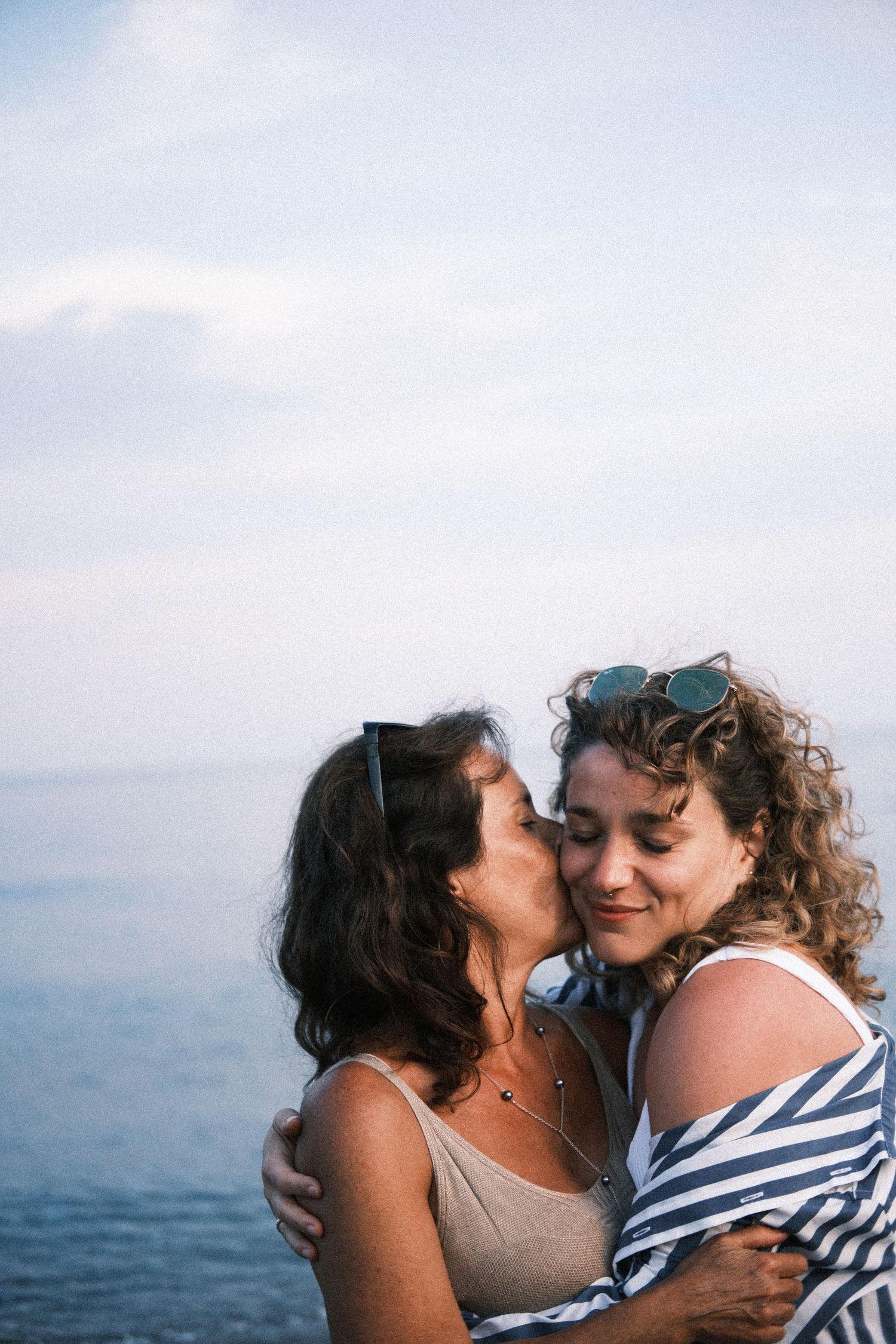 A couple shares tender moments by the ocean, wearing casual summer attire against a misty coastal backdrop.