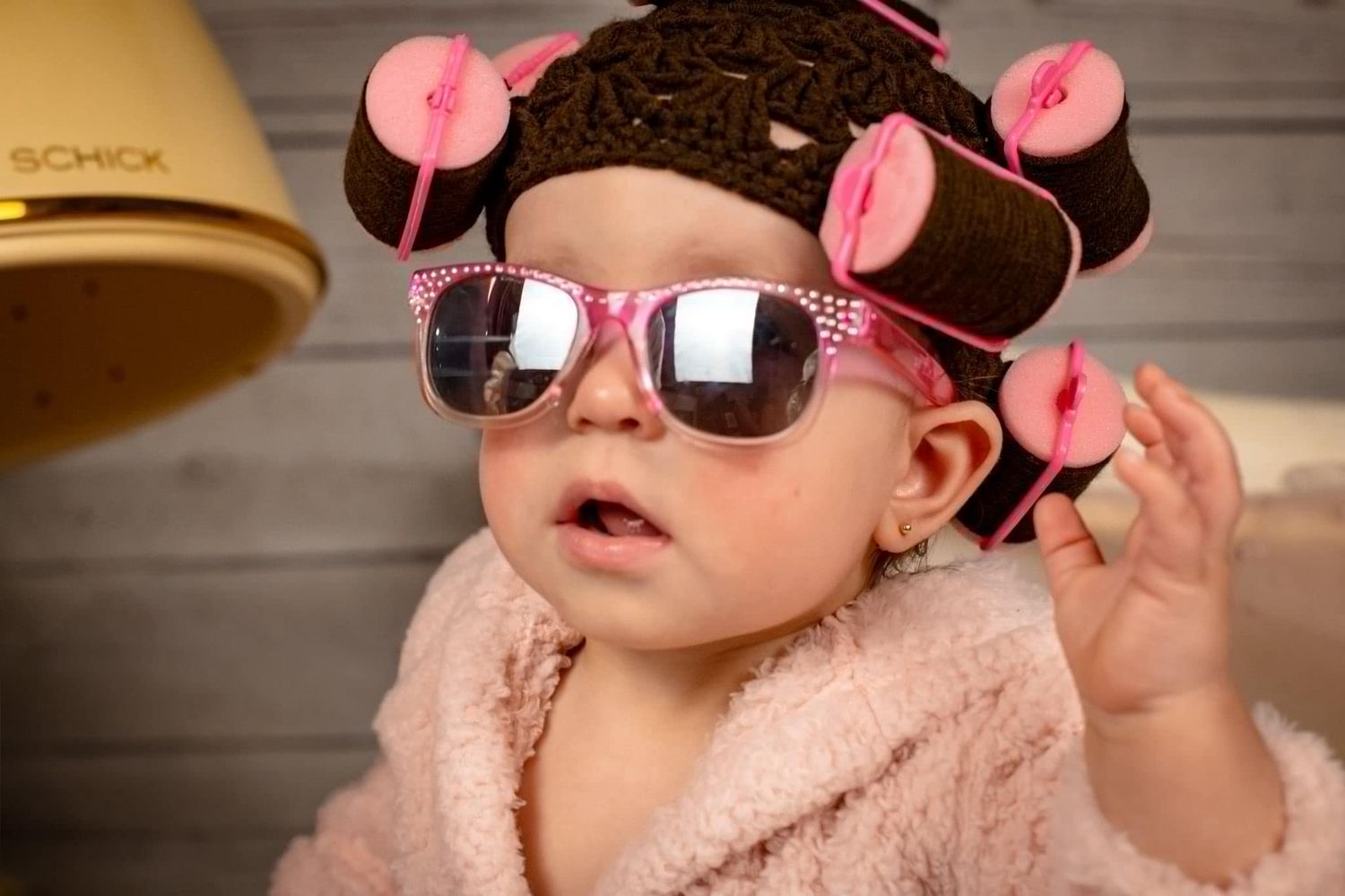 Baby wearing sunglasses and pink hair curlers poses in a fuzzy pink robe.