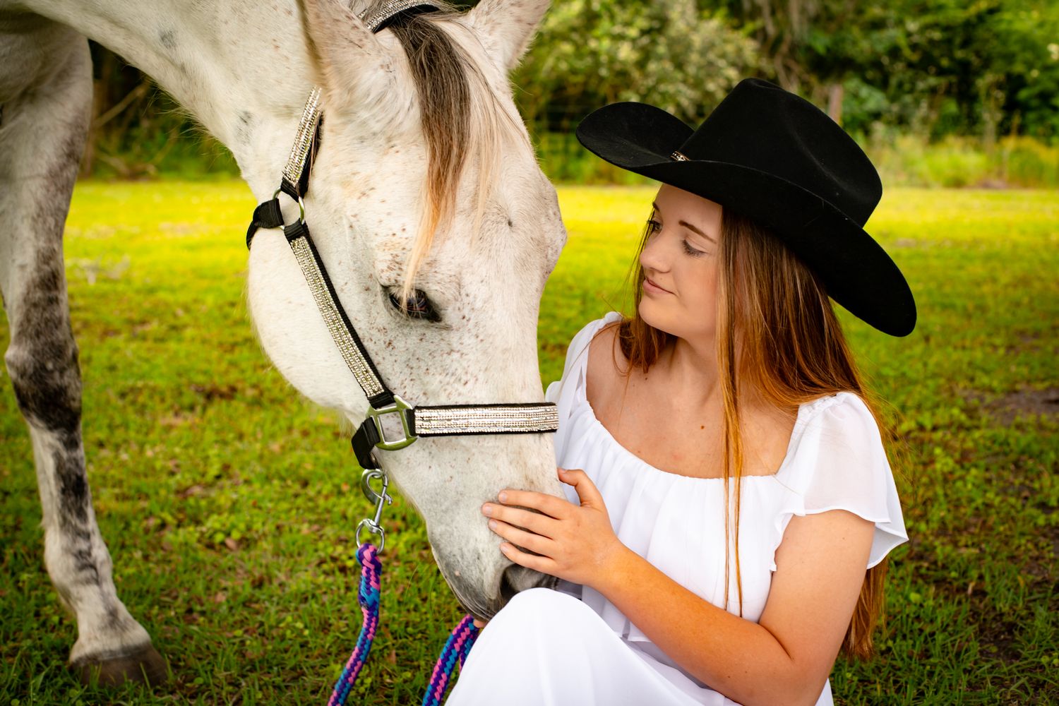 A person in a white top and cowboy hat pets a white horse in a sunny grassy field.
