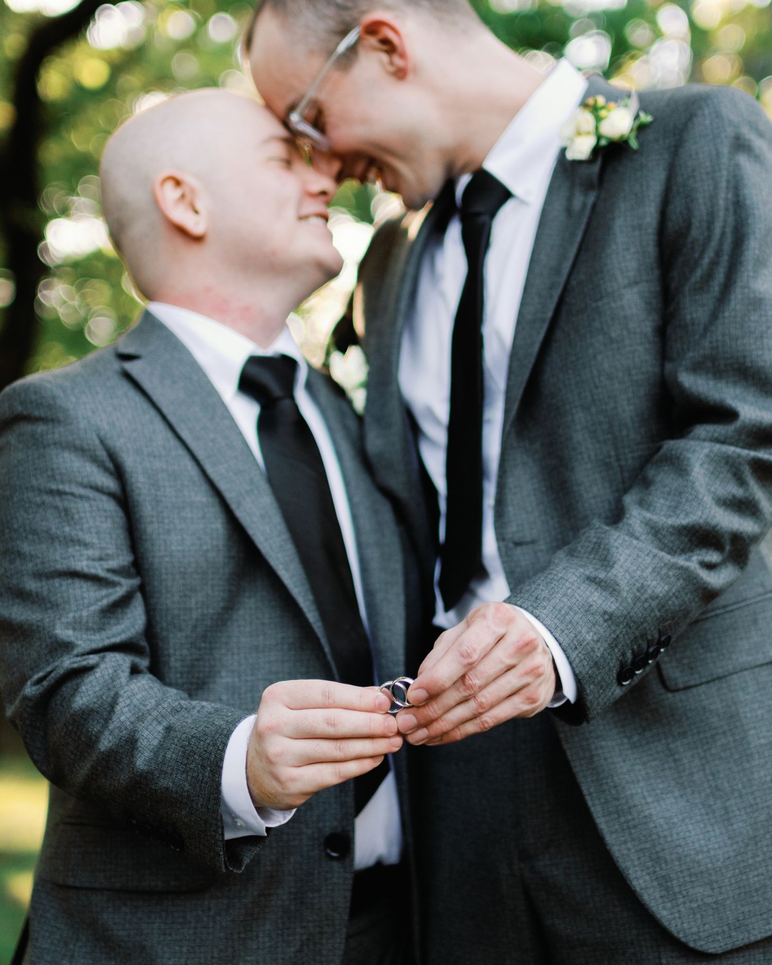 Two people in gray suits and black ties share an intimate moment outdoors with bokeh lighting in the background.