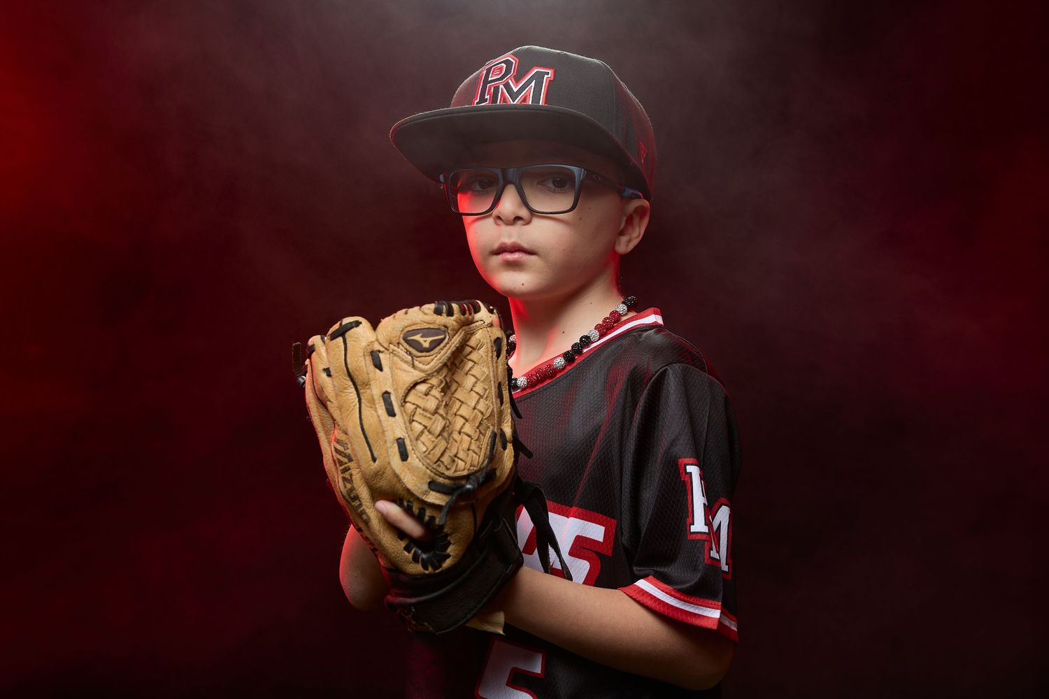 A young baseball player in a black jersey and PM cap poses with their glove in a dramatic dark studio portrait series.