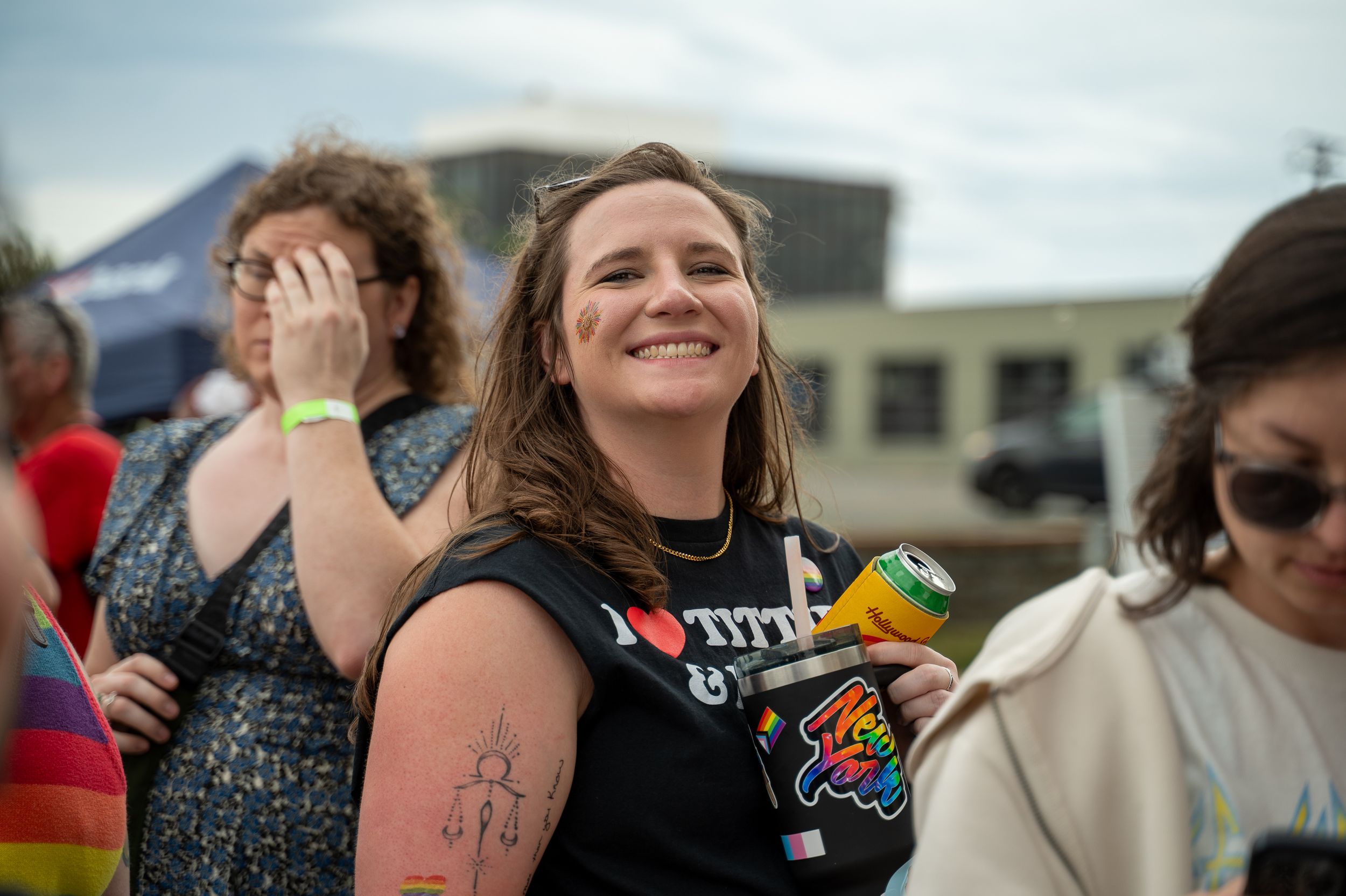 Smiling woman in black tank top holding a drink at an outdoor Pride celebration event.