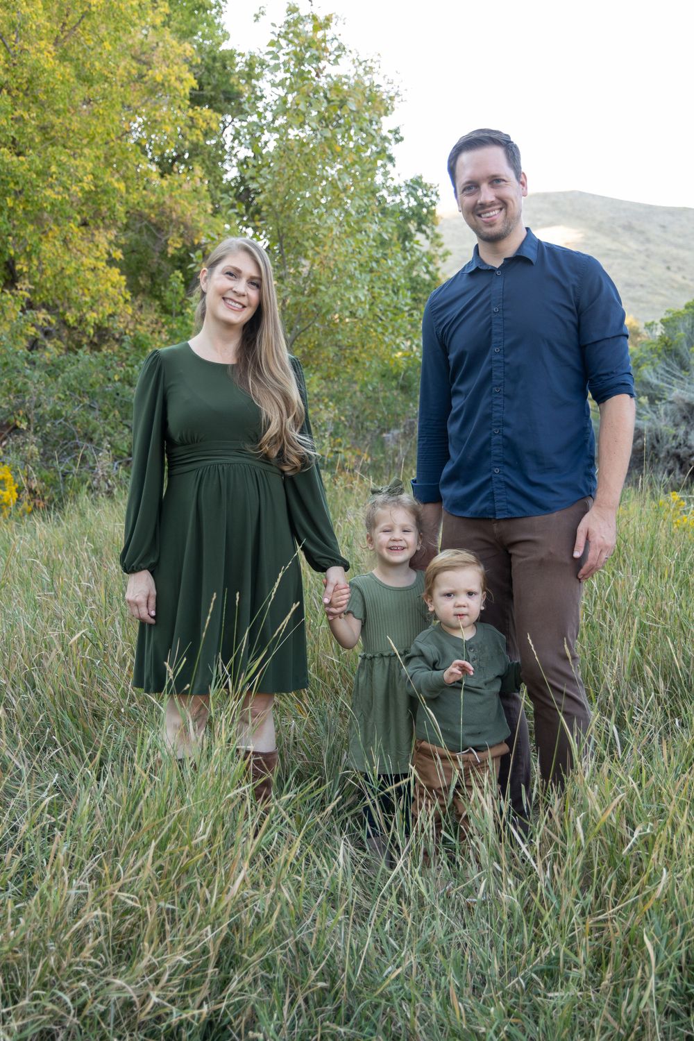 A group in coordinating green and blue outfits poses together in tall grass against a mountain backdrop.