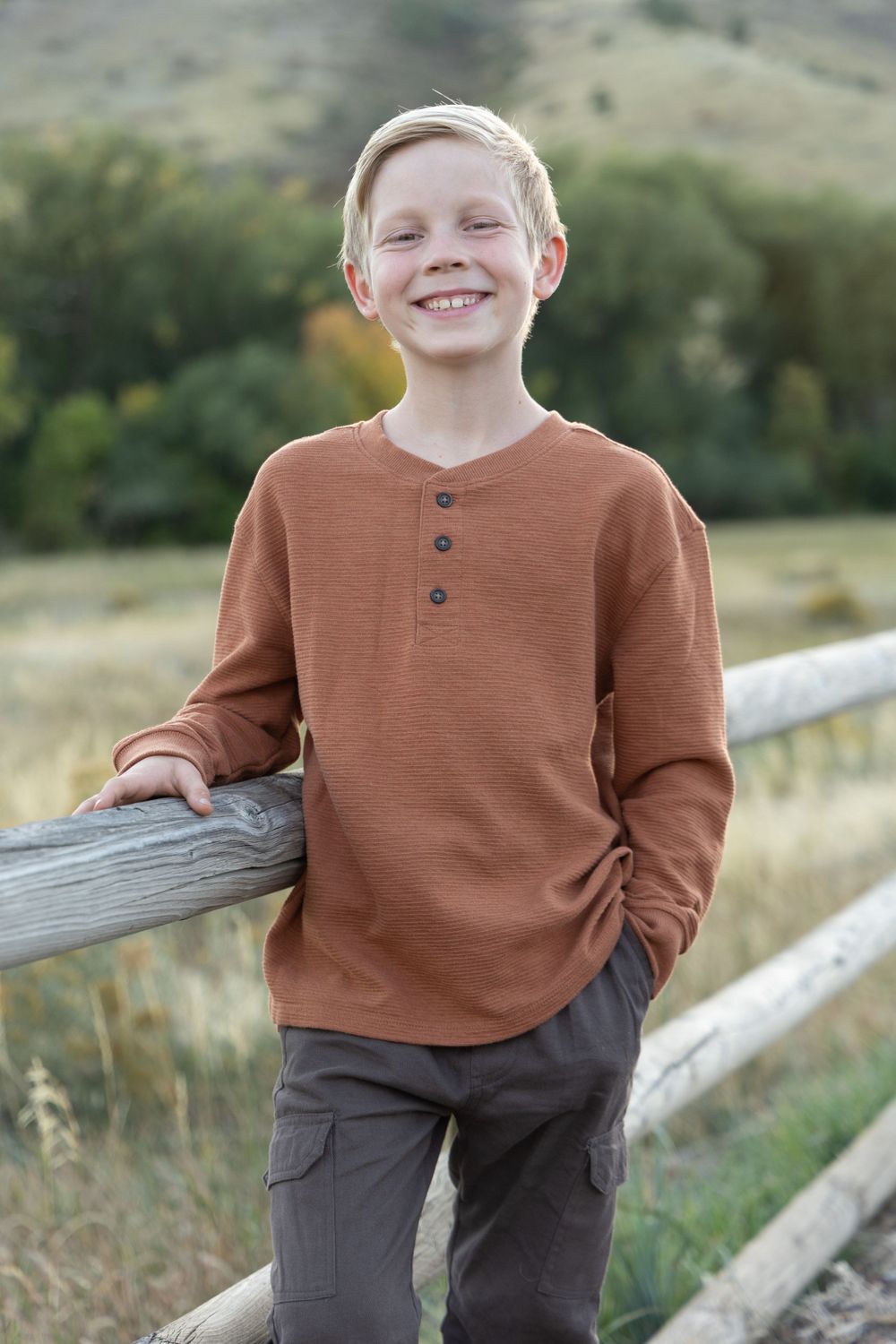 Portrait in rustic setting wearing brown henley shirt while leaning on wooden fence with hills in background.