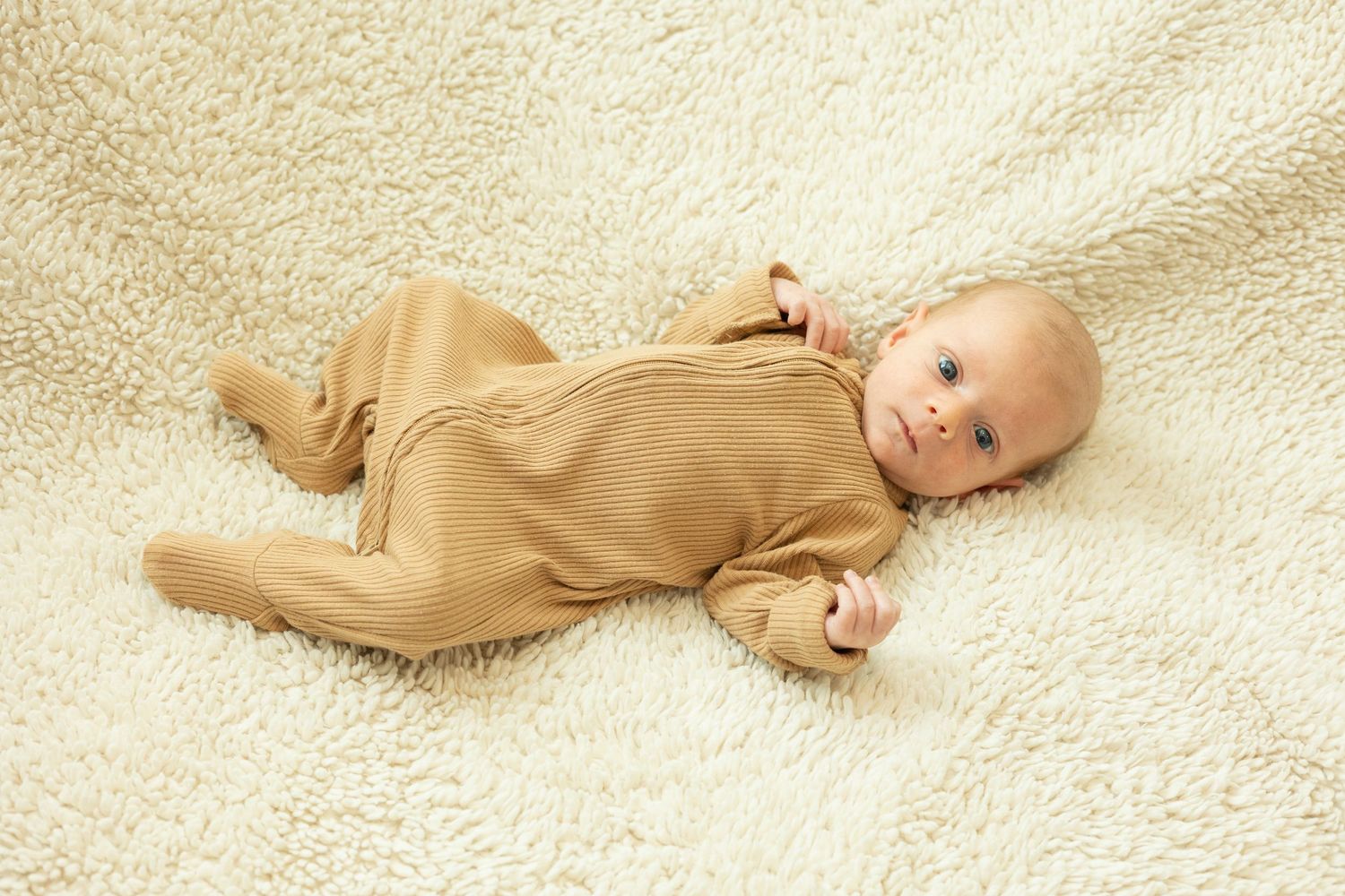 Baby in tan ribbed onesie lying on cream textured blanket.