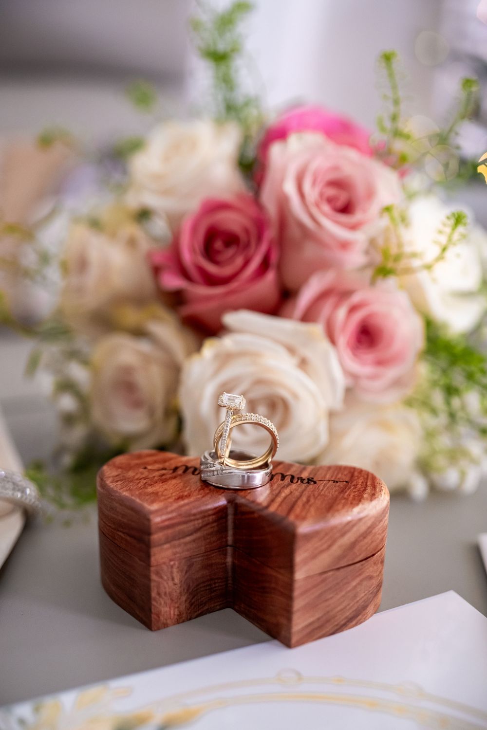 Wedding rings on wooden heart-shaped box with pink and white roses in background.