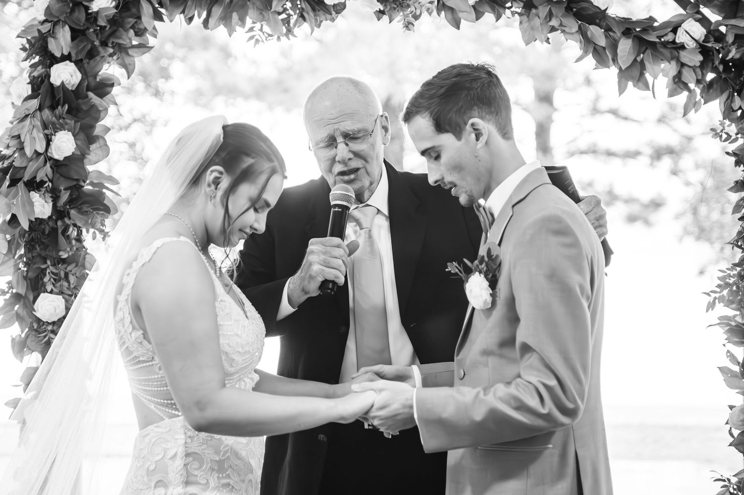 Black and white wedding ceremony photo showing couple exchanging rings under floral arch.