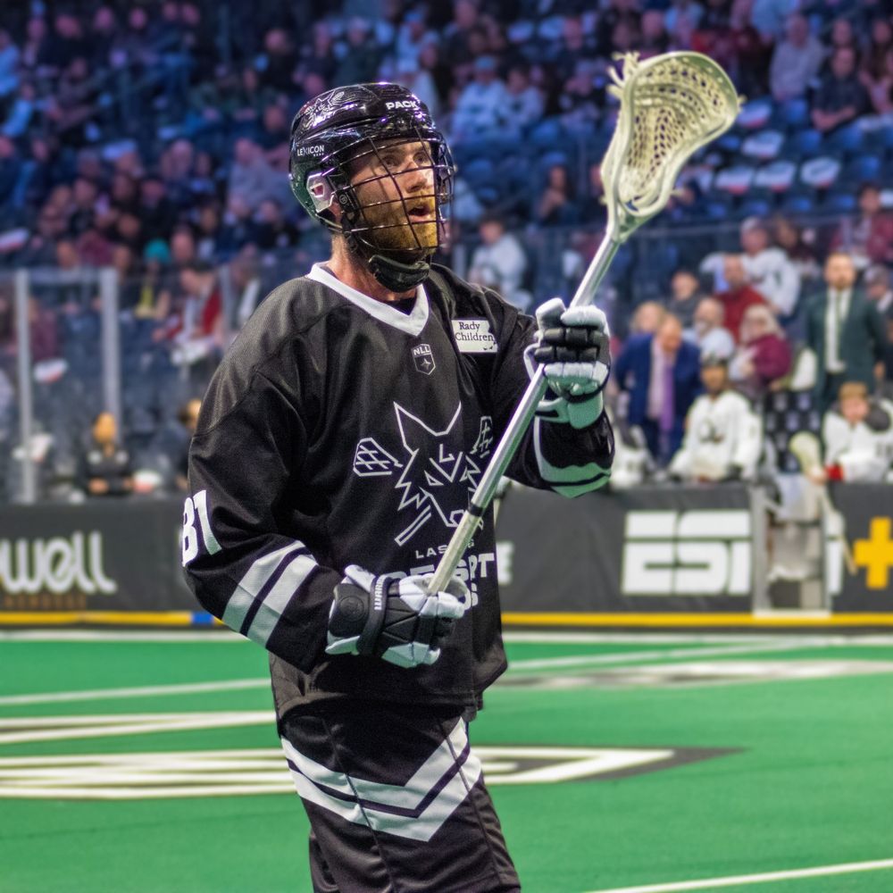 Lacrosse player in black uniform holding stick ready to pass during indoor NLL game.