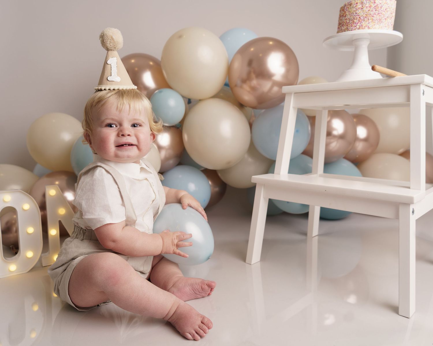 First birthday photo setup with neutral balloons, white furniture, and party decorations.