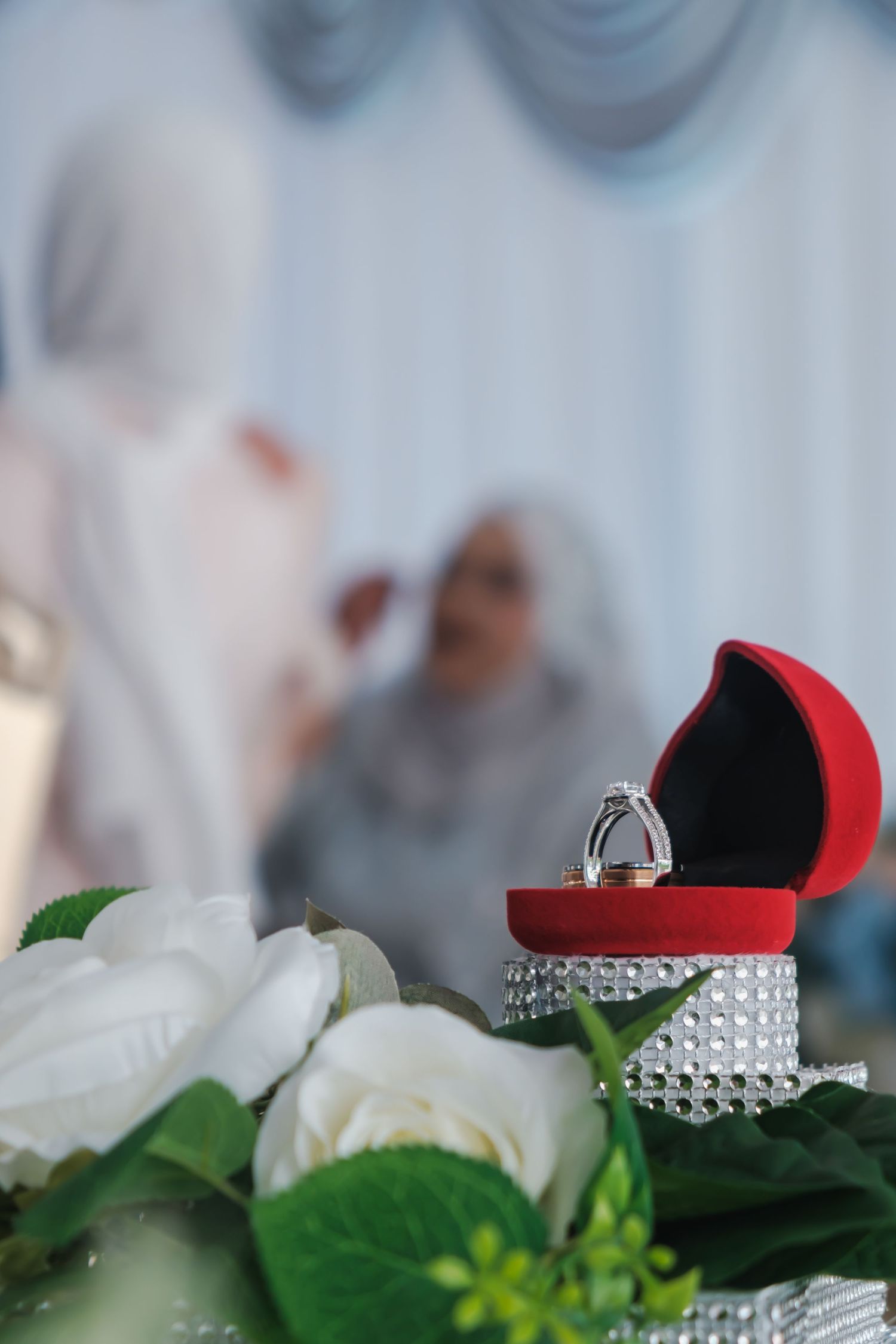 Diamond engagement ring in a red velvet box displayed with white roses and crystal decorations.