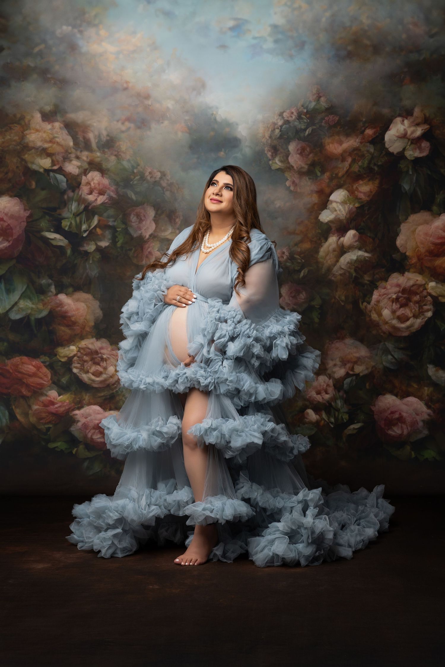 A dreamy maternity portrait with a flowing powder blue tulle gown against a dark floral backdrop with roses.
