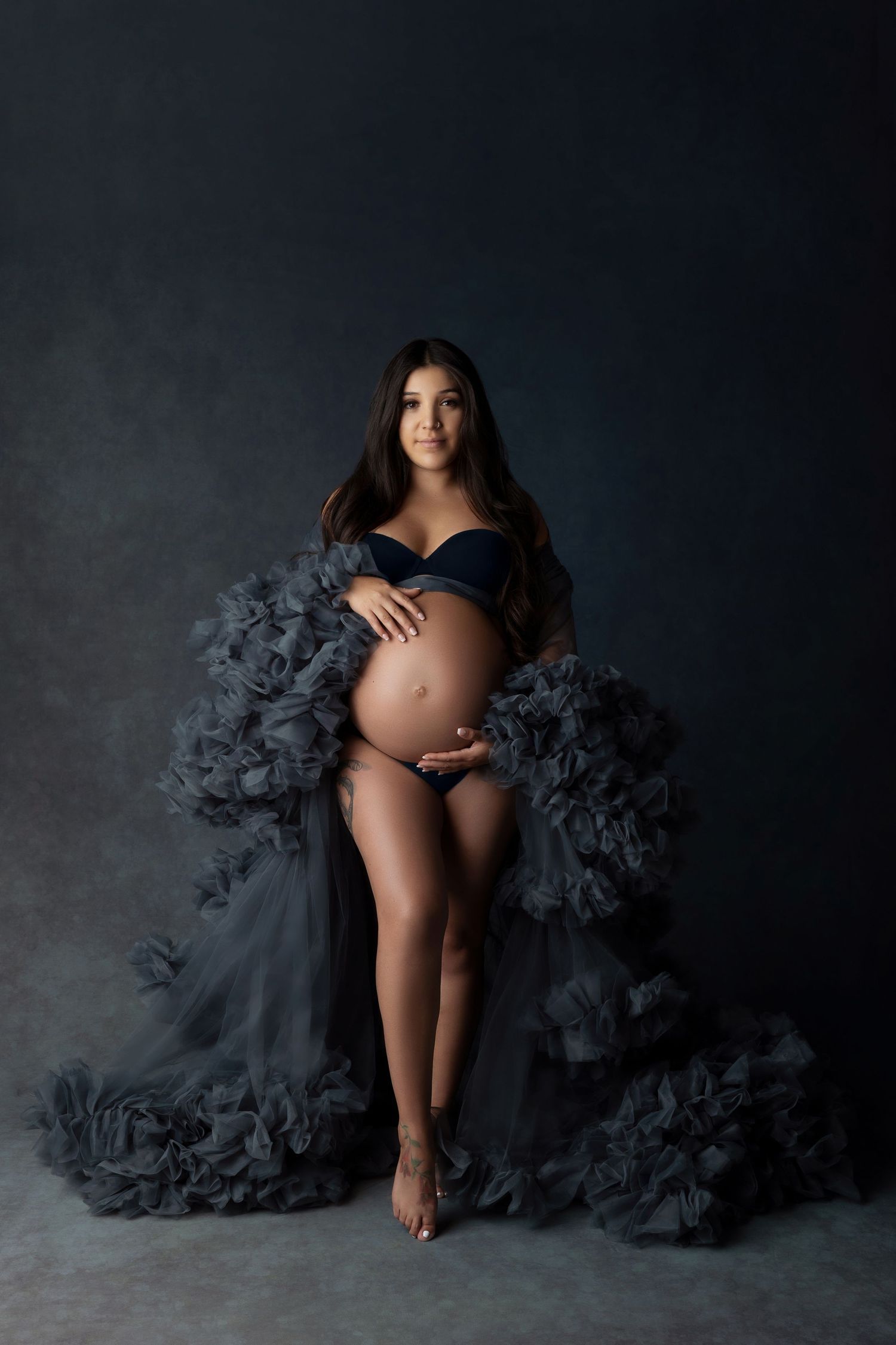 Artistic maternity portrait featuring black lingerie and dramatic gray tulle against dark backdrop.