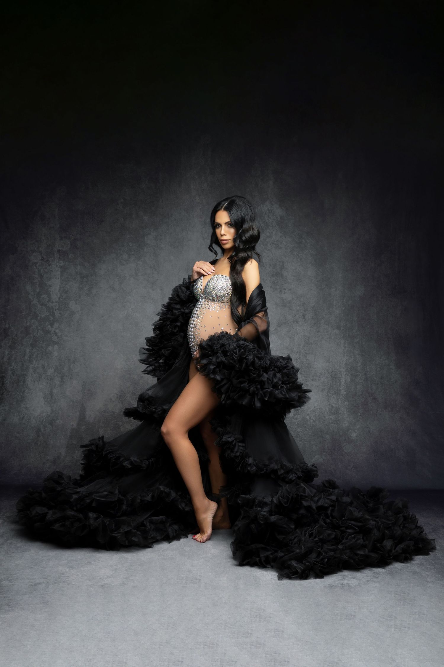 Dramatic maternity photo with flowing black ruffled dress and silver embellished top against dark moody backdrop.