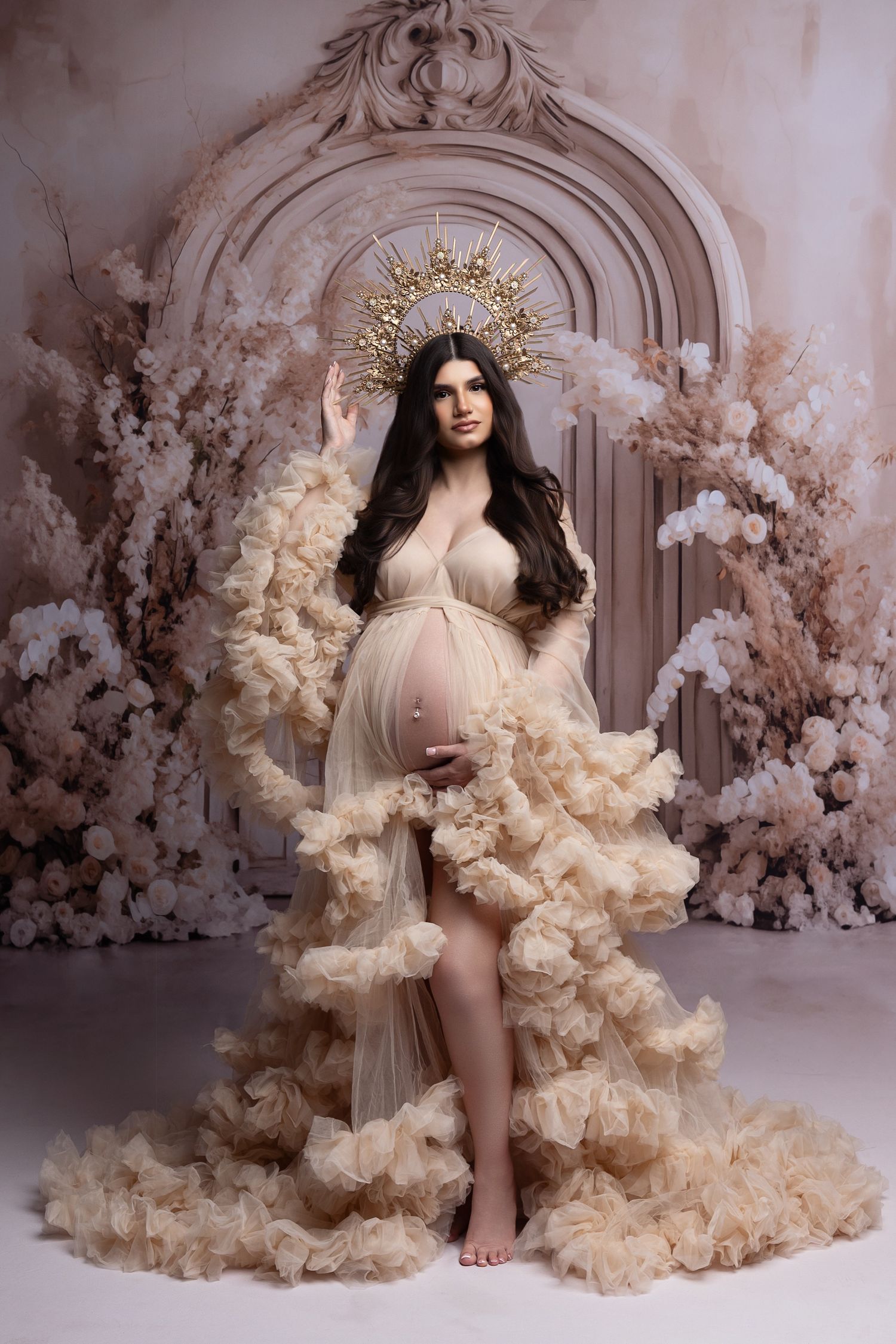 Pregnant woman in gold crown and ruffled champagne gown poses gracefully among white flowers in elegant maternity photo.