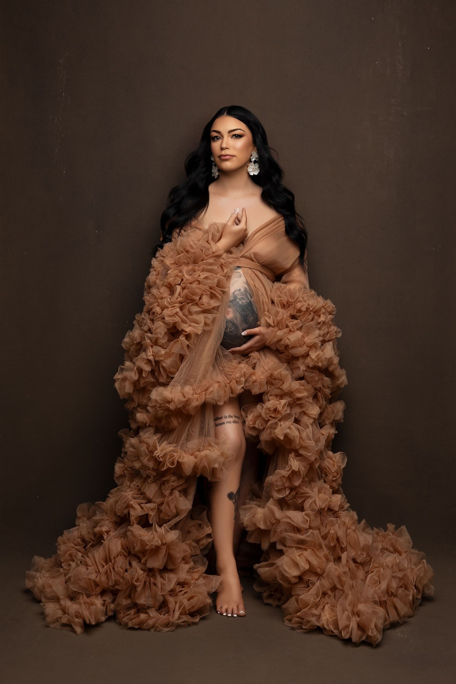 A figure poses in a dramatic brown ruffled tulle gown against a dark brown studio backdrop.