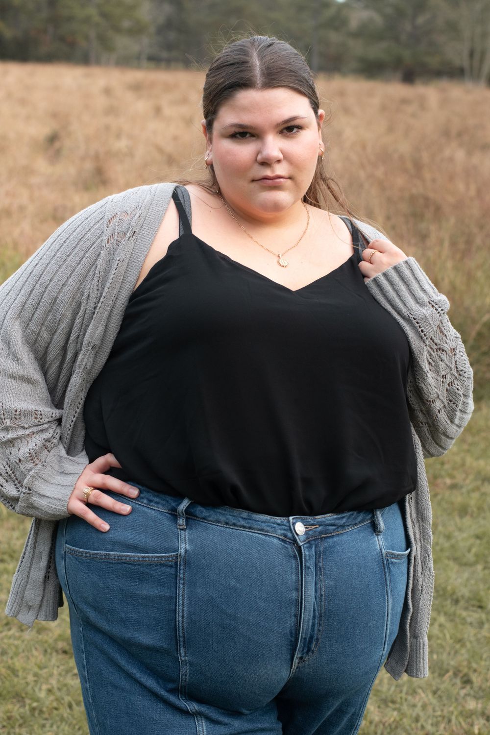 A person in a gray cardigan and black top poses confidently in a field.