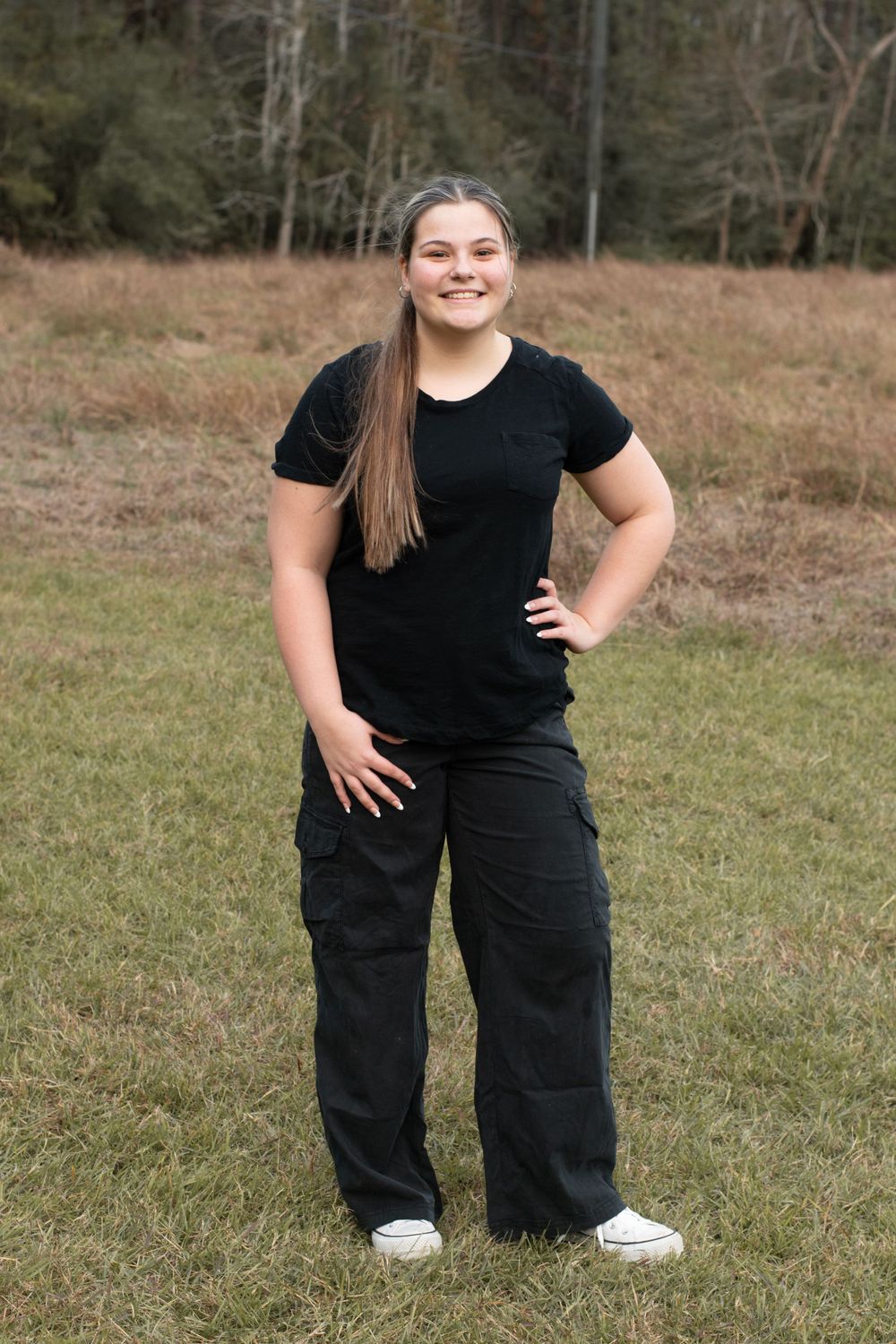 A person in black clothing poses confidently with hand on hip in an outdoor field setting.