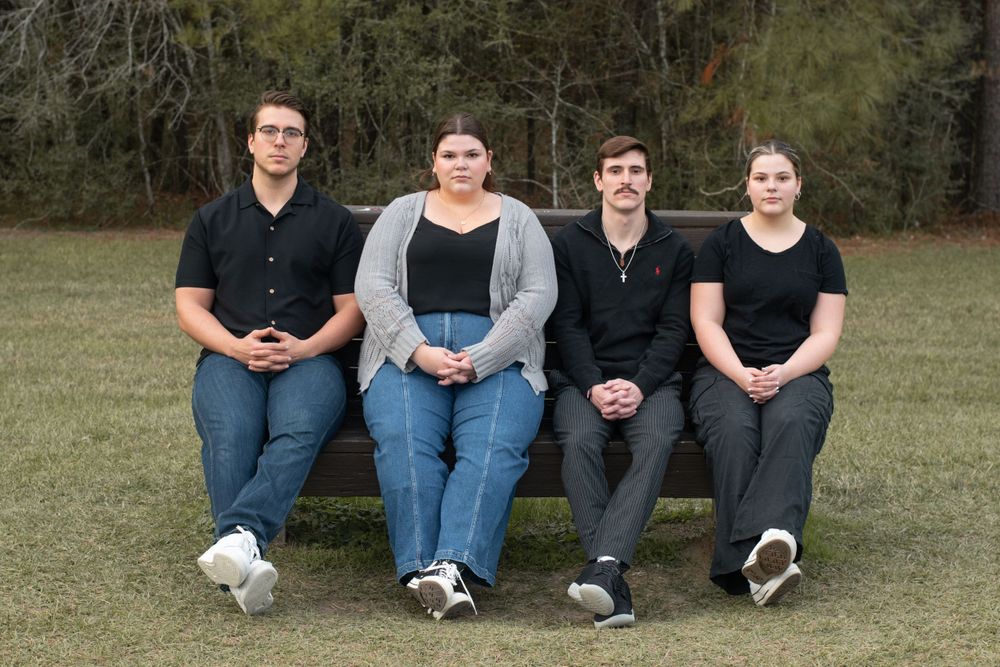 Four people sitting together on a bench in casual attire for an outdoor group photo.