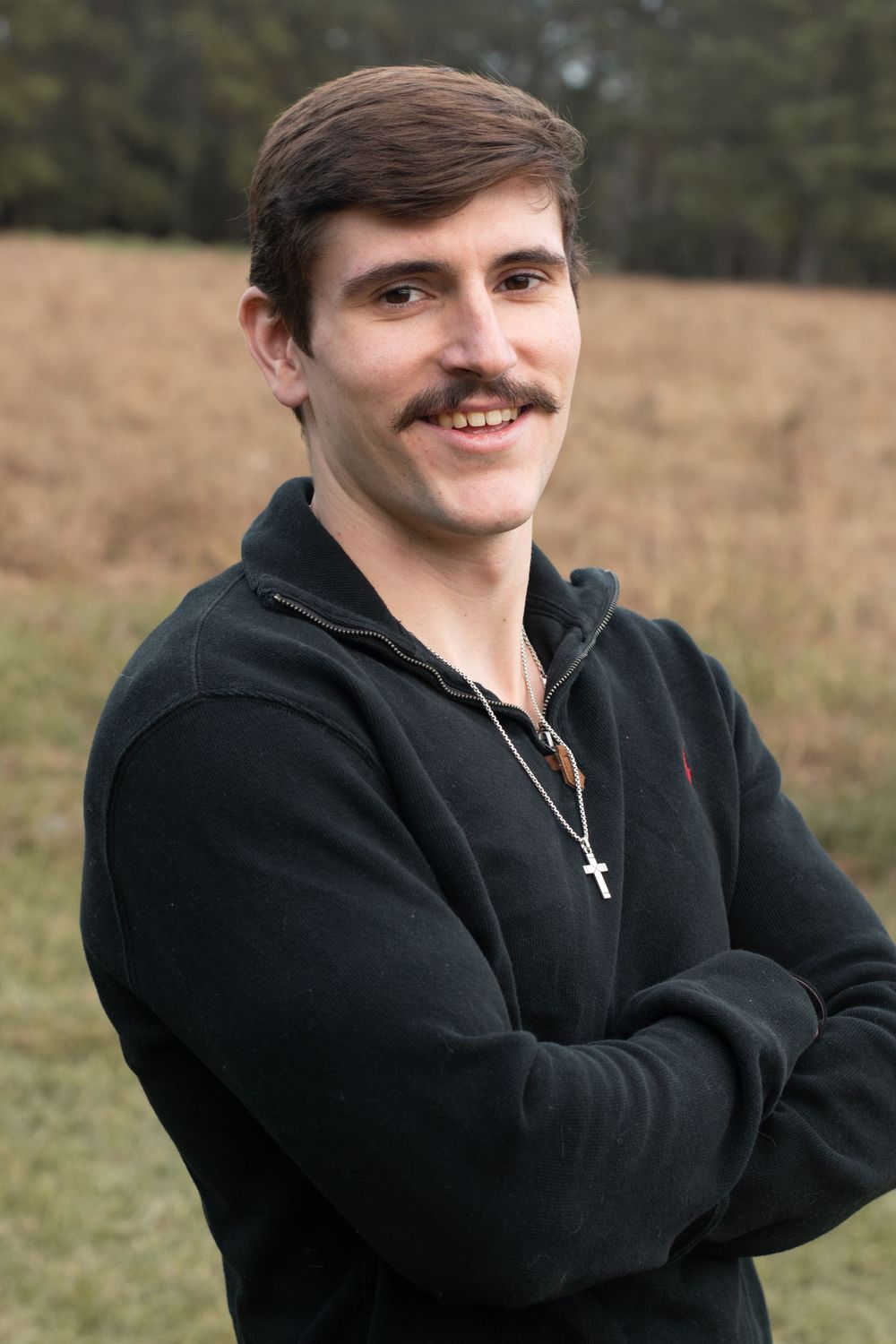 A person in a black sweater with a cross necklace poses against a natural outdoor backdrop.