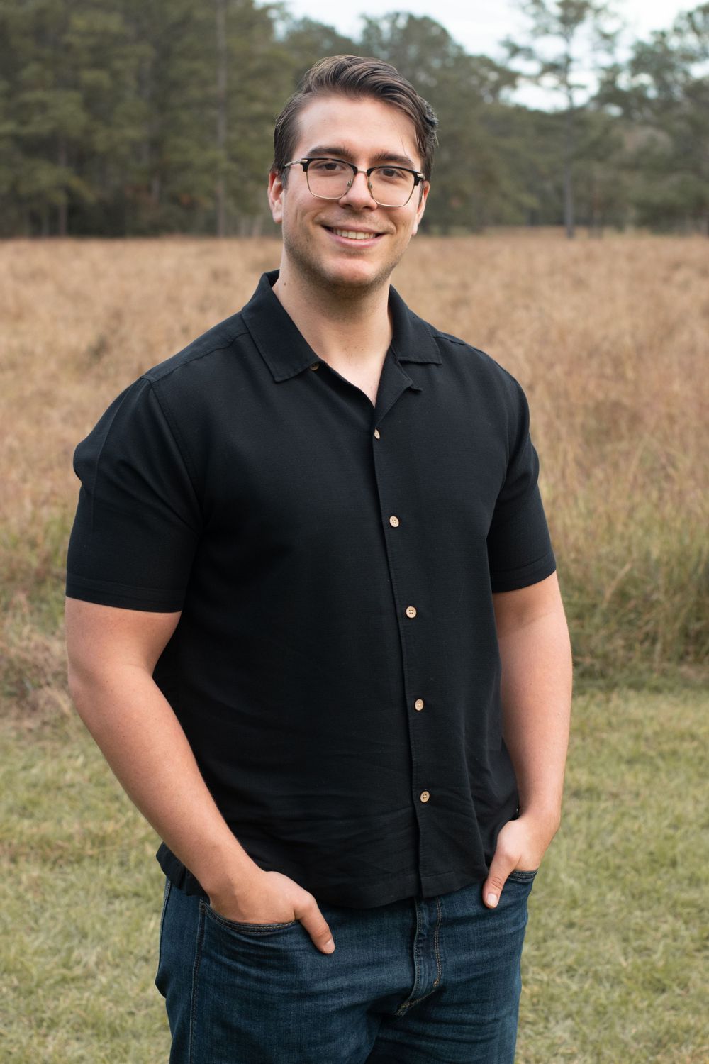 A person in a black button-down shirt smiles warmly in an outdoor portrait setting.