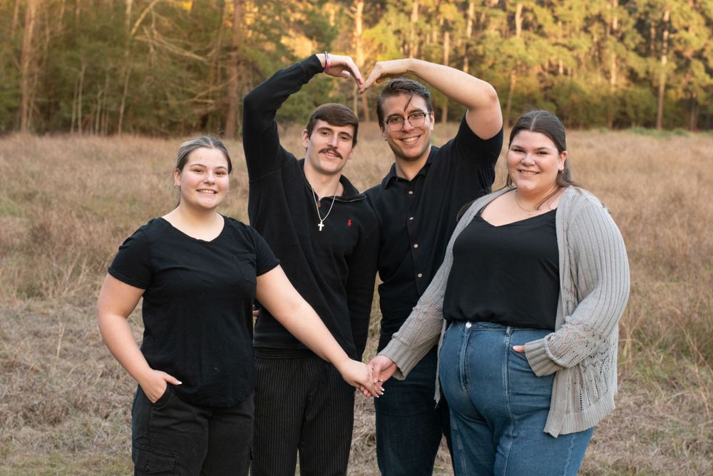 A group of four people in black outfits pose together in a grassy field at sunset.