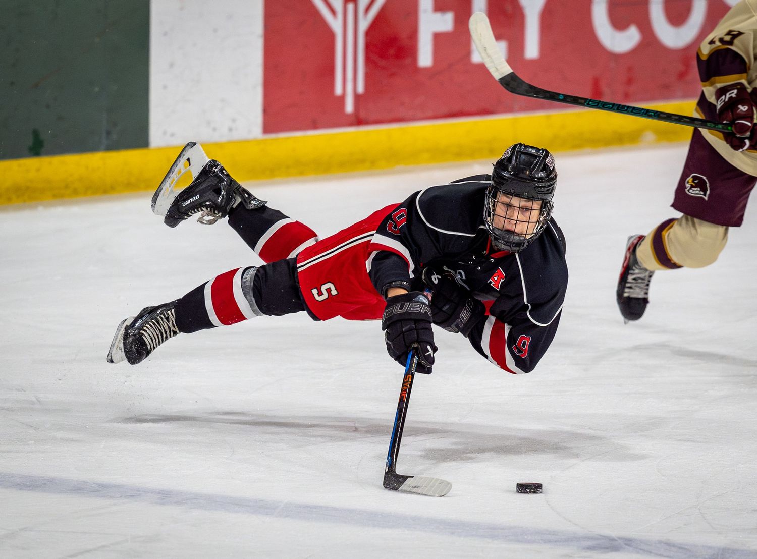 Hockey player in black jersey makes dramatic diving play while reaching for puck on ice.