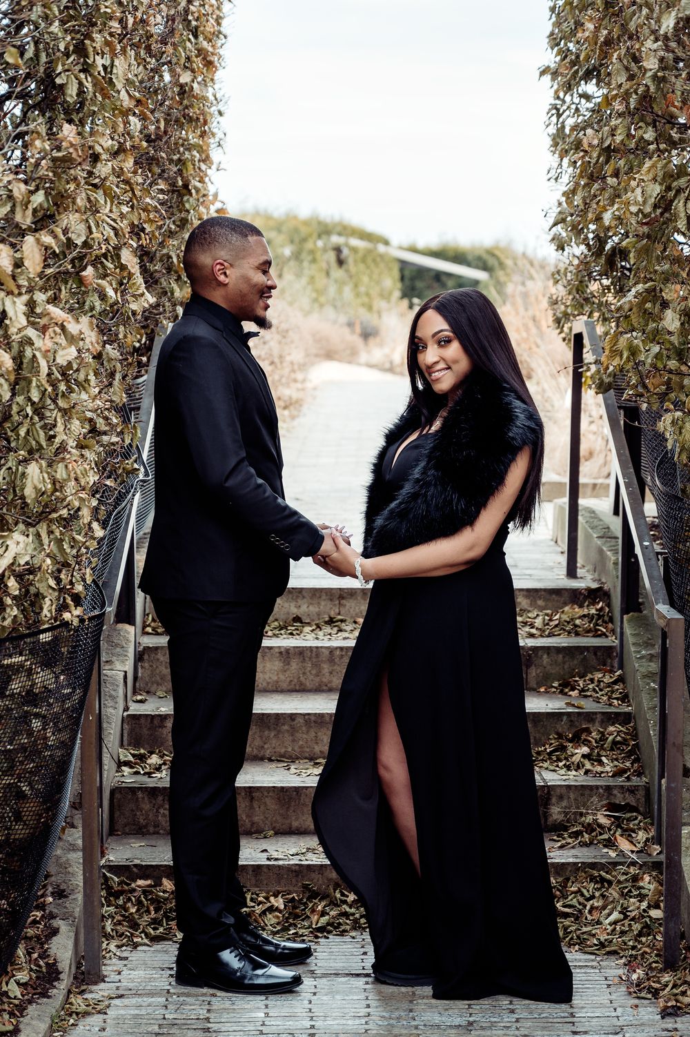 Couple in formal black attire holding hands on stone steps surrounded by dried vines.