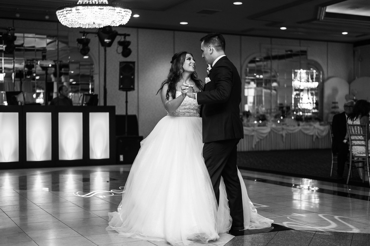 A couple shares their first dance in a reception hall with a glamorous chandelier overhead.