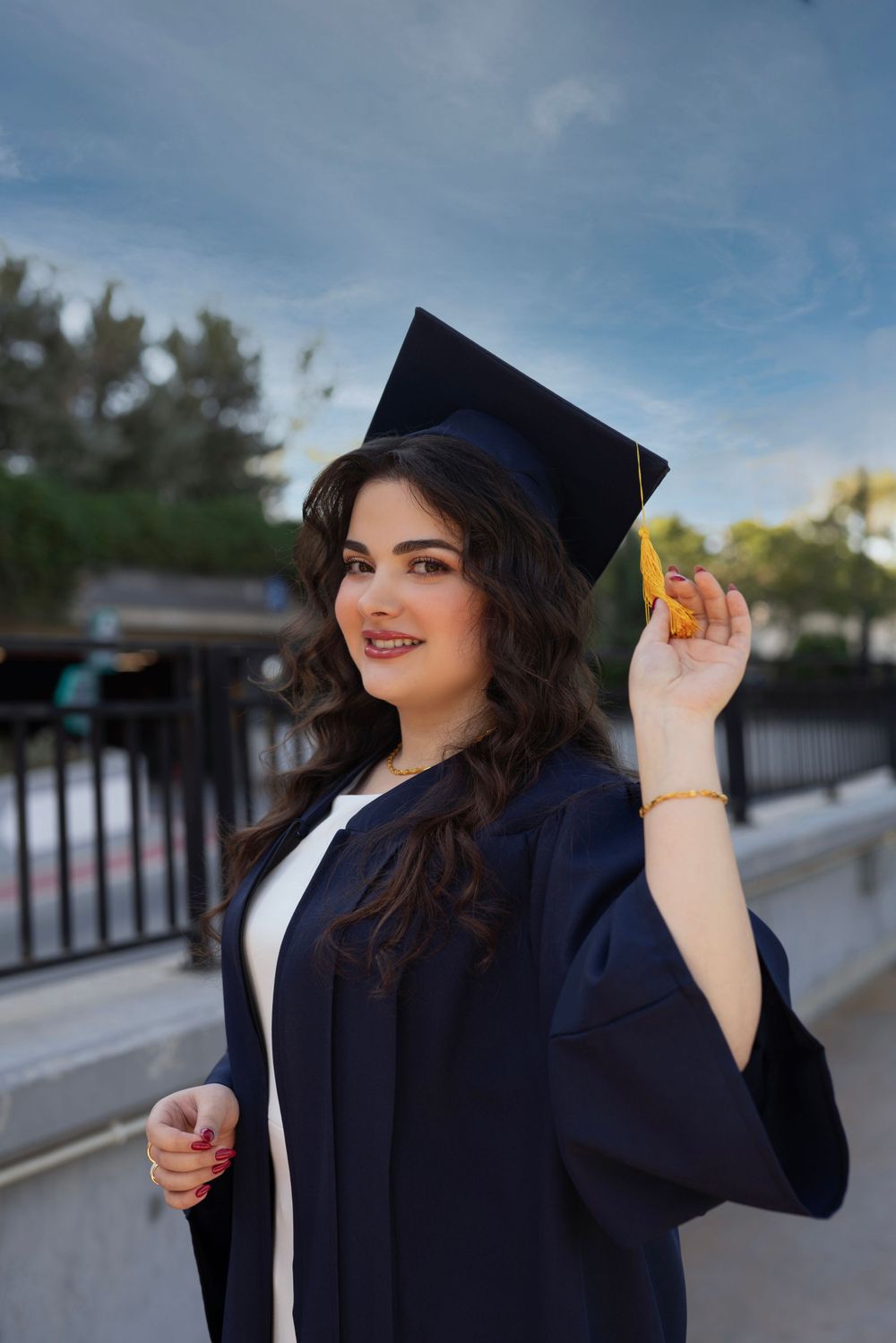 A graduate in navy blue cap and gown poses outdoors on a sunny day with a blue sky background.