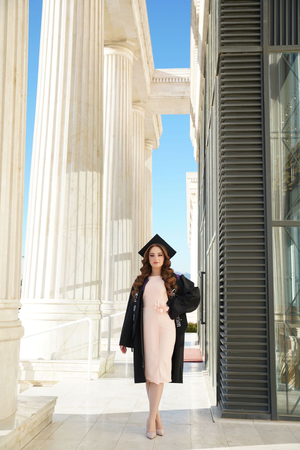 Graduate in cap and gown stands elegantly between white classical columns on a sunny graduation day.