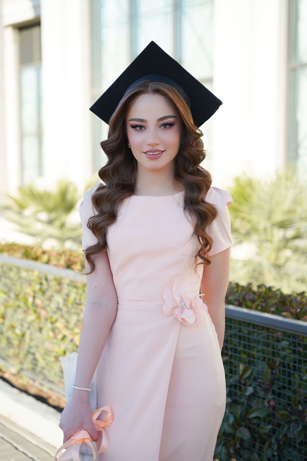 Graduate in soft pink dress and mortarboard cap poses outdoors with elegant curled hairstyle.