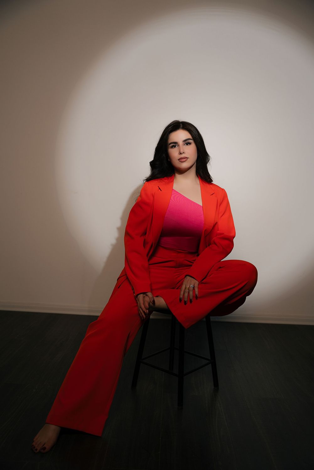 A person in a vibrant red pantsuit and pink top poses on a stool against a neutral wall with dramatic lighting.