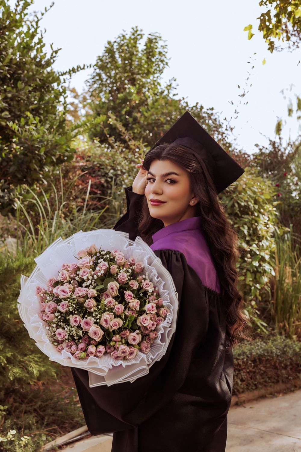 A graduate in black cap and gown holds a beautiful bouquet of pink roses during an outdoor graduation photoshoot.