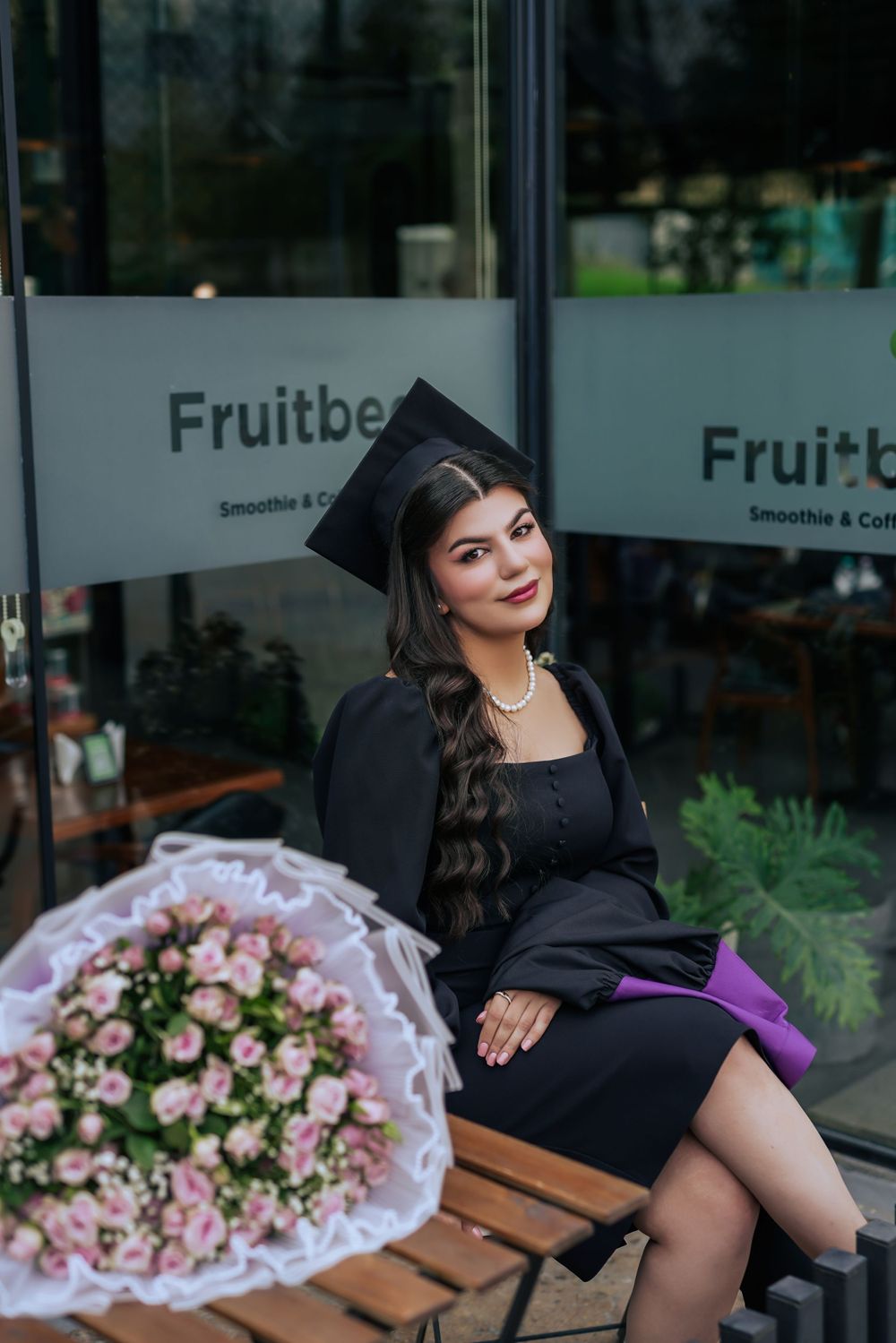 Graduate in black cap and gown sits on bench with pink floral bouquet outside Fruitbe storefront.