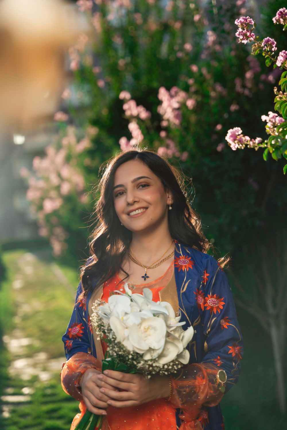 A woman in an orange dress and blue floral shawl holds white flowers while standing near blooming pink blossoms.